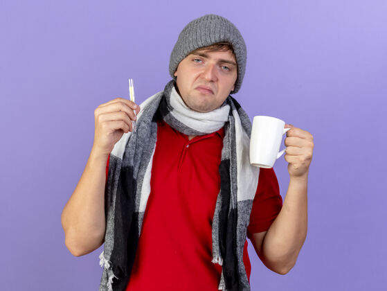 衣服悲伤的年轻英俊的金发病男子戴着冬天的帽子和围巾拿着温度计和杯子看着前面孤立的紫色墙壁悲伤人脸
