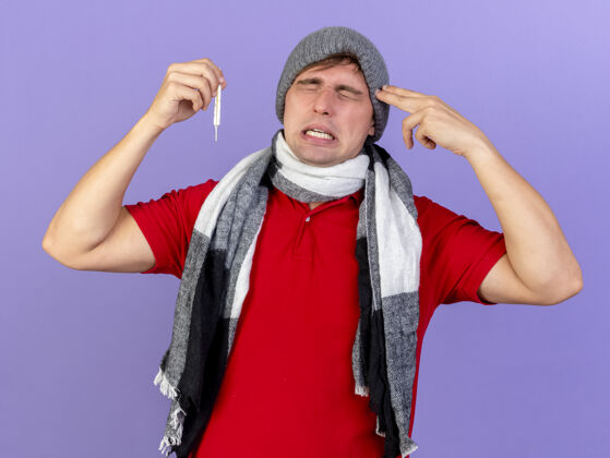 人受够了年轻英俊的金发病患戴着冬天的帽子和围巾拿着温度计做手势闭着眼睛孤立在紫色的墙上摆姿势闭上围巾
