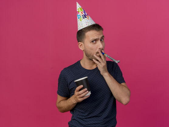 人戴着生日帽的自信帅哥拿着纸杯吹口哨孤立地贴在粉色的墙上 留着复印空间吹帽子口哨