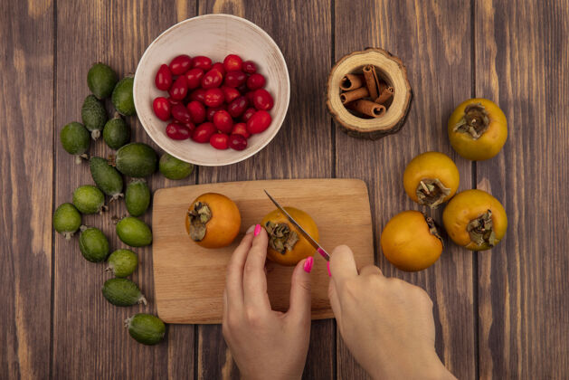 新鲜女性手在木制厨房板上切柿子水果的俯视图 刀子上放着山茱萸樱桃 碗里放着新鲜的柿子和飞珠 隔离在木制背景上水果手食物