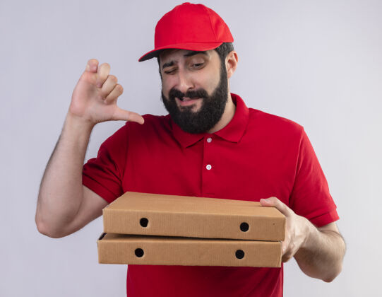 披萨年轻英俊的白人送货员穿着红色制服 戴着帽子 看着披萨盒 在白色背景上孤立地竖起大拇指拇指帅气帽子