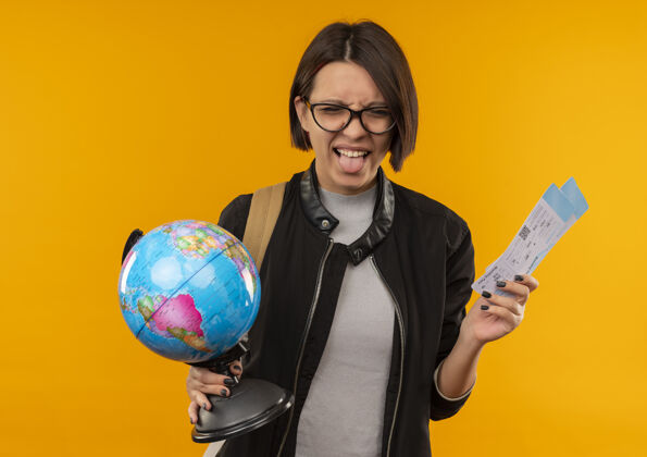 女孩顽皮的年轻学生女孩戴着眼镜 背着包 手里拿着飞机票 地球仪上的舌头被隔离在橙色的背景上机票显示穿