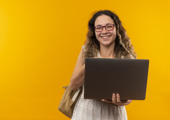 眼镜快乐的年轻漂亮的女学生戴着眼镜 背着包拿着笔记本电脑隔离在黄色快乐黄色背