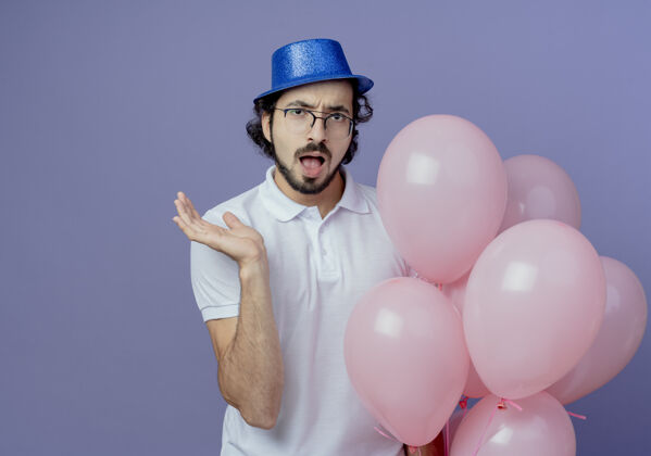 手惊讶的帅哥戴着眼镜 戴着蓝色帽子 手里拿着气球 摊开手孤立在紫色的背景上惊喜气球戴着