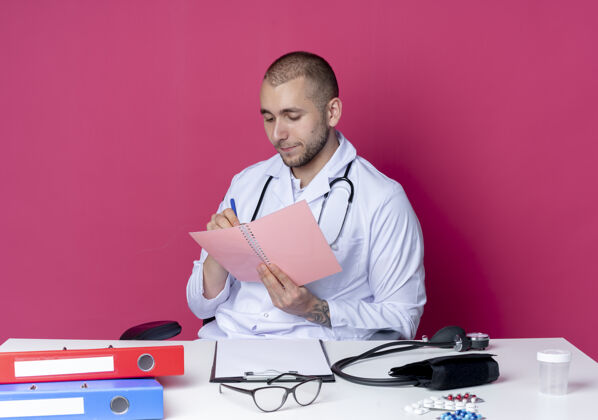 听诊器年轻的男医生穿着医用长袍 手持听诊器坐在办公桌旁 手里拿着工作工具 看着便笺簿 用粉色背景上孤立的笔在上面写着什么工作医疗拿着