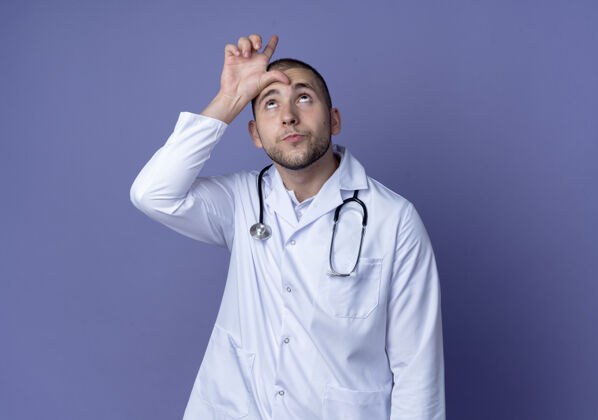 听诊器年轻的男医生穿着医用长袍 戴着听诊器 在紫色的背景上做着一个孤立的手势 还有一个复制空间医疗男性穿