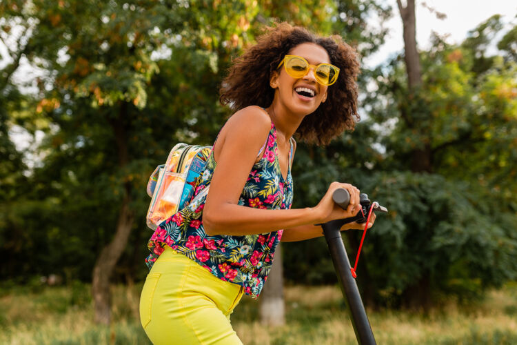 女士年轻时尚的黑人女子在公园里玩得很开心骑着电动脚踏车在夏天的时尚风格 五颜六色的时髦服装 穿着背包和黄色太阳镜道路多彩人