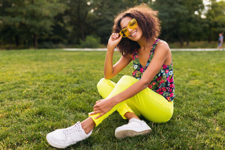卷曲年轻时尚的黑人女子在公园里尽情享受夏日的时尚风格 五颜六色的时髦装扮 坐在草地上戴着黄色的太阳镜和裤子 穿着运动鞋人魅力休闲