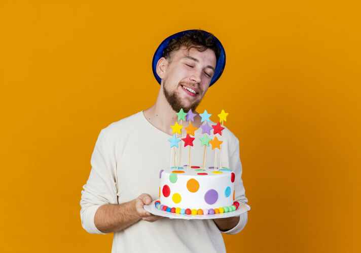 蛋糕微笑的年轻帅哥斯拉夫党人戴着党帽举行生日蛋糕与星星孤立在橙色背景与复制空间抱着帽子斯拉夫人