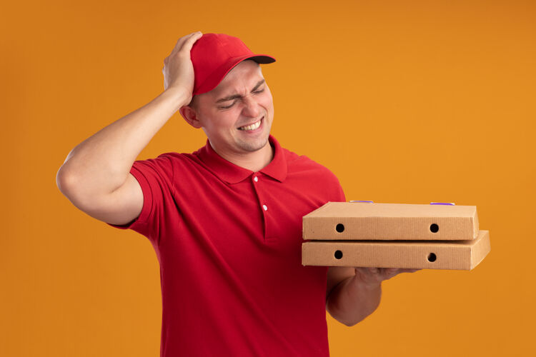 制服遗憾的是 年轻的送货员穿着制服 戴着帽子 看着披萨盒 手放在隔离在橙色墙上的头上年轻人人头男人
