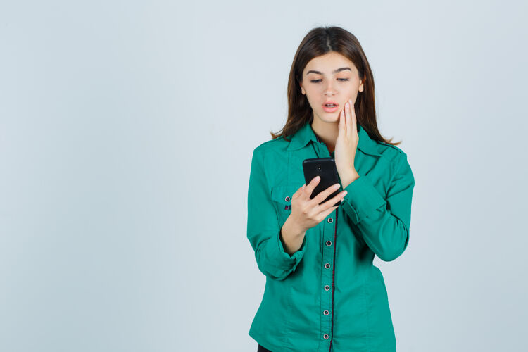 电话年轻的女孩在看手机短信 手放在脸颊上 穿着绿色上衣 黑色裤子 神情专注 俯视前方年轻客厅前面