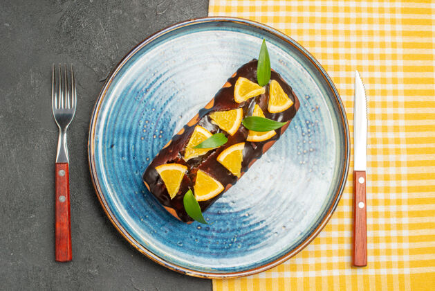 服务用橘子和巧克力装饰的美味蛋糕 用叉子和刀子放在黑桌子上晚餐深色陶瓷器皿