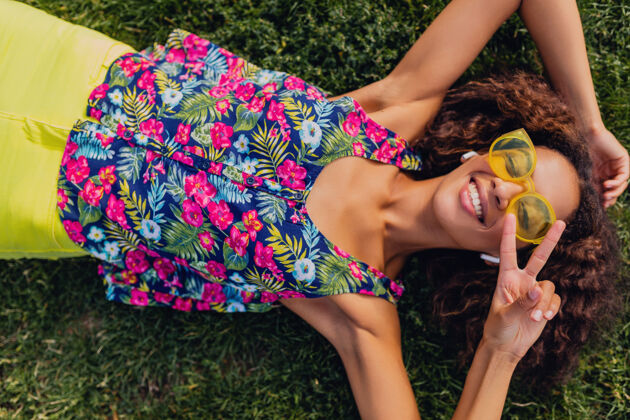 肖像年轻时尚的黑人女子戴着无线耳机听音乐在公园里玩得很开心 夏日时尚风格 五颜六色的时髦装扮 躺在草地上 俯瞰风景积极年轻卷发