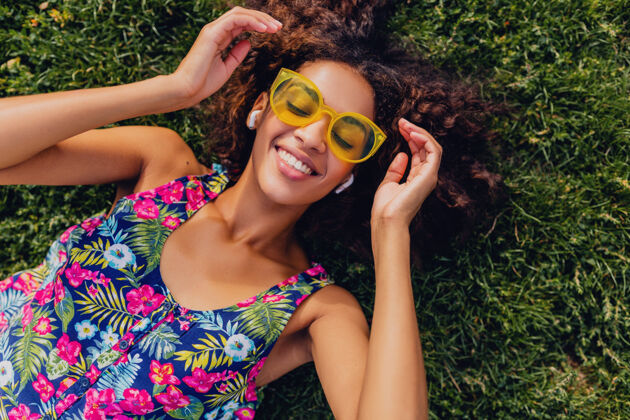 黄色年轻时尚的黑人女子 戴着无线耳机听音乐 躺在公园的草地上玩得很开心 夏日的时尚风格 五颜六色的时髦装扮 俯瞰风景耳机微笑女人