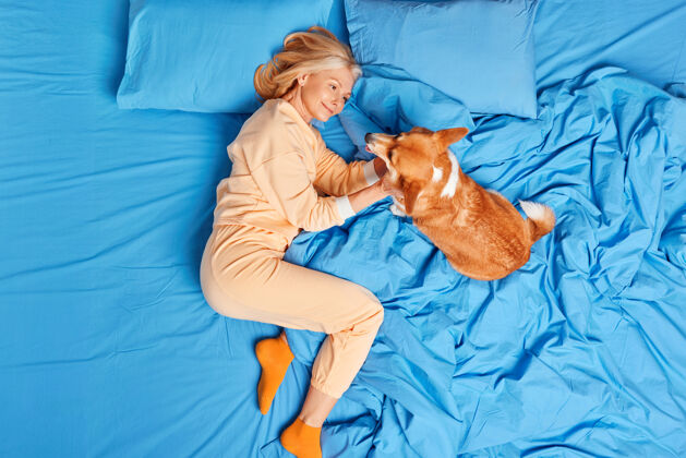 放松穿着睡衣的欧洲老年妇女在卧室里和心爱的宠物玩耍 一起躺在床上 享受美好的一天中年女性作为家庭的一员 表达对狗的爱和关怀床友谊枕头
