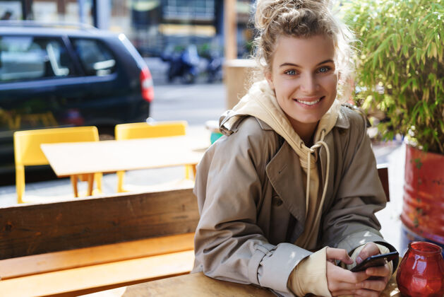 手机坐在咖啡馆户外桌子上的美丽微笑的女孩在咖啡馆里用手机的女人微笑等待桌子