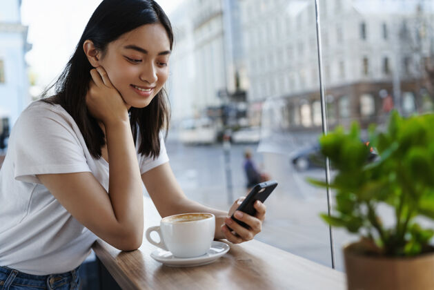 杯子在靠近窗户的餐厅里 一个女孩一边用智能手机聊天 一边喝咖啡 一边对着手机屏幕微笑城市年轻人桌子