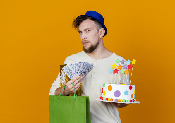 年轻困惑的年轻帅哥斯拉夫党家伙戴着党帽子拿着礼品盒钱纸袋和生日蛋糕与明星看着相机孤立的橙色背景与复制空间空间钱盒子
