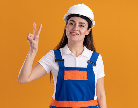 和平身着制服的年轻建筑工人微笑着 在橙色的墙上展示着和平的姿态微笑建筑工人制服