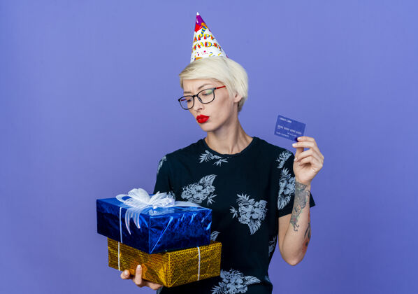 紫色年轻的金发派对女孩戴着眼镜 戴着生日帽 手里拿着礼品盒和信用卡 看着紫色背景上有复制空间的盒子年轻盒子持有