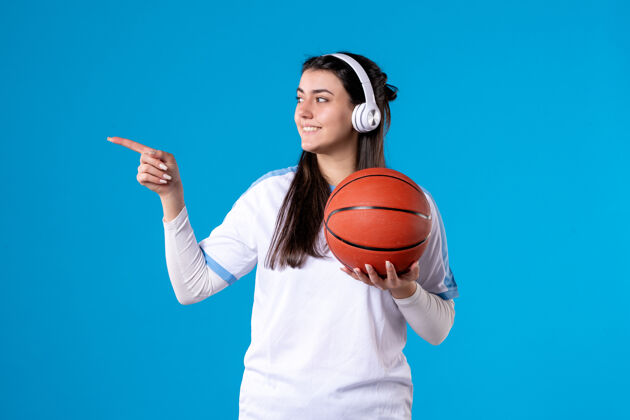篮球正面图：戴着耳机拿着篮球的年轻女性站在蓝色的墙上人青年前面