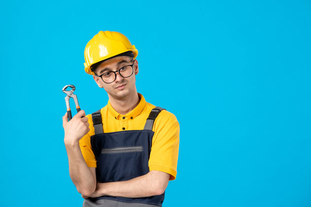 工人蓝色墙上穿着制服 戴着钳子的头盔的男建筑工人的正面图杂工专业男人