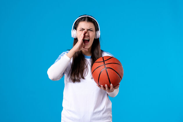 竞争正面图：戴着耳机的年轻女性拿着篮球站在蓝色的墙上耳机尖叫年轻女性