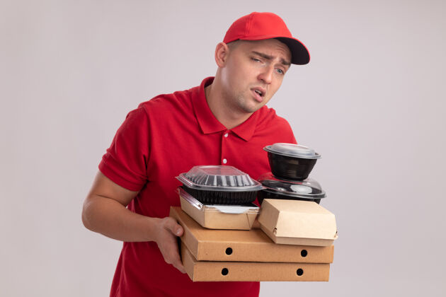 男人疲惫的年轻送货员穿着制服 戴着帽子 把食物容器放在隔离在白墙上的比萨饼盒上穿披萨容器