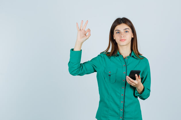 手机手持手机的年轻女士的肖像 在绿色衬衫中显示ok手势 看起来很高兴的正面视图黑发女孩女性