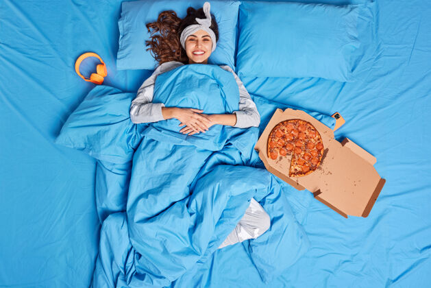 蓝色快乐的黑发年轻女子在舒适的床上享受慵懒的一天戴着头巾躺在柔软的毯子下吃着美味的比萨饼微笑房间女性