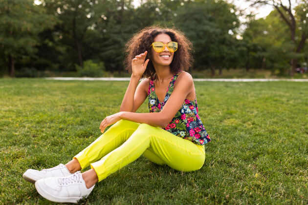 时尚年轻时尚的黑人女子在公园里尽情享受夏日的时尚风格 五颜六色的时髦装扮 坐在草地上戴着黄色的太阳镜和裤子 穿着运动鞋积极黑色女孩