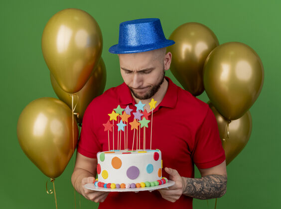 站着年轻帅气的斯拉夫派对小伙子戴着派对帽站在气球前拿着生日蛋糕看着绿色的背景绿色抱着小伙子