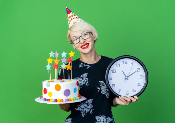 眼镜微笑的年轻金发派对女孩戴着眼镜 戴着生日帽 拿着生日蛋糕和时钟 看着隔离在绿色背景和复制空间的相机金发时钟年轻