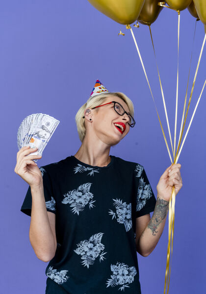 金发快乐的年轻金发派对女孩戴着眼镜和生日帽拿着气球和钱看着孤立在紫色背景上的气球快乐紫色年轻