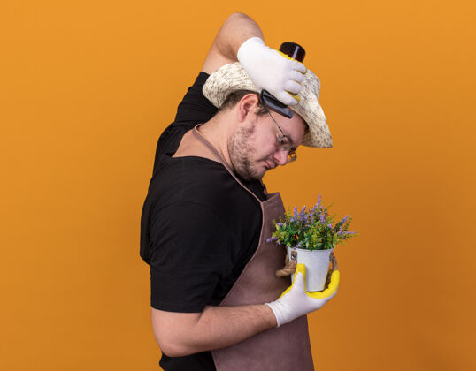 瓶站在剖面图年轻的男园丁戴着园艺帽和手套在花盆里浇花喷雾瓶隔离在橙色的墙上手套浇水穿