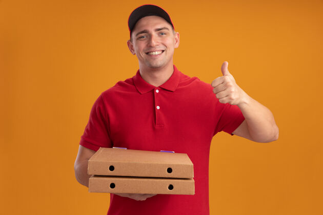 盒子微笑的年轻送货员穿着制服 戴着帽子 拿着披萨盒 在橙色的墙上孤立地竖起大拇指送货披萨帽子