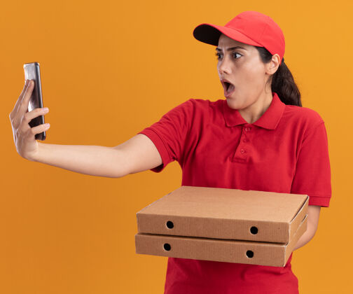 盒子惊讶的年轻送货女孩穿着制服 戴着帽子拿着披萨盒 独自在橙色的墙上自拍拿着惊喜自拍