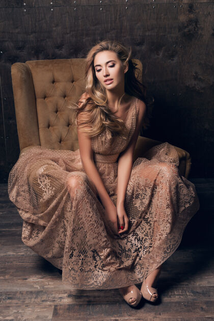 衣服穿着闪亮蕾丝晚礼服的美丽性感模特坐在米色的椅子上美丽坐着身体