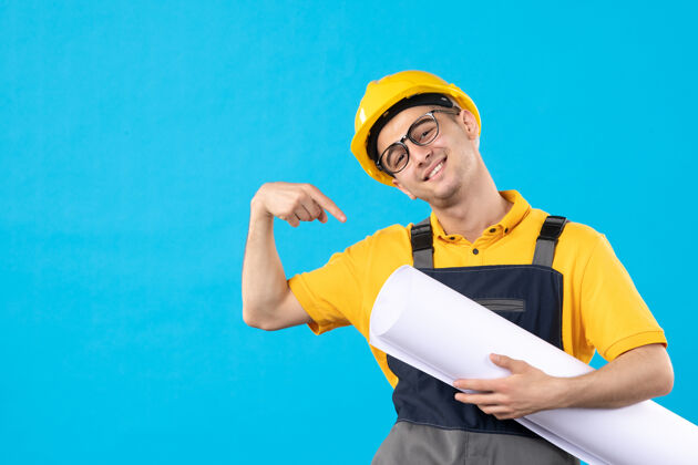 肖像身穿黄色制服和头盔的男性建筑工人正视图 蓝色墙上有平面图计划工作男性