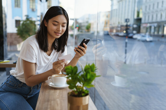 雇用亚洲现代女孩在咖啡馆里 喝咖啡 在智能手机里输入信用年轻人柜台信用卡