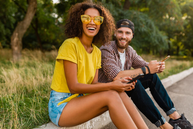兴奋快乐的年轻微笑的朋友坐在公园里用智能手机 男人和女人一起玩时尚多彩享受