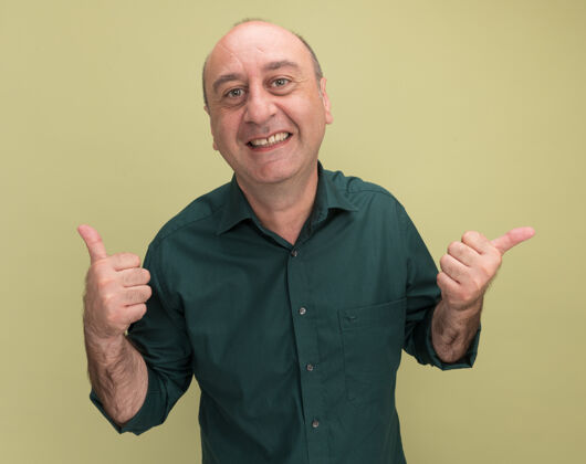 微笑微笑的中年男子身穿绿色t恤 大拇指朝上 指着橄榄绿的墙壁上有复制空间的隔离侧穿橄榄点