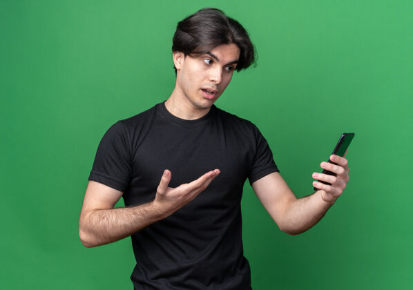 电话迷茫的年轻帅哥穿着黑色t恤拿着手机指着隔离在绿色墙上的手机年轻拿着家伙