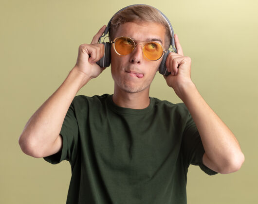 橄榄令人印象深刻的看边年轻帅哥穿着绿色衬衫戴着眼镜和耳机隔离在橄榄绿的墙上耳机壁板穿