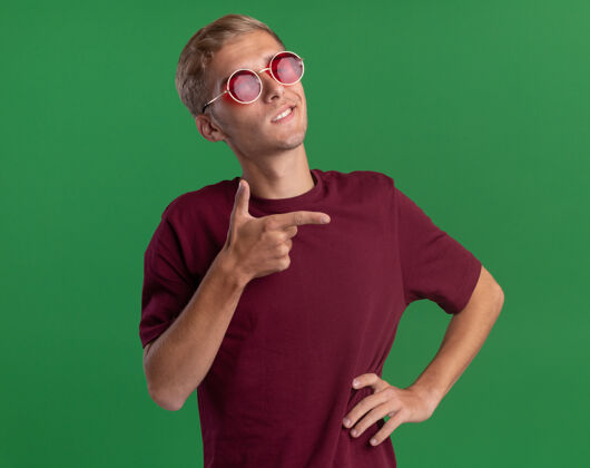年轻自信的年轻帅哥穿着红衬衫 戴着眼镜 手放在臀部 指着隔离在绿色墙上的一侧壁板臀部家伙