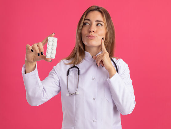 穿着年轻的女医生穿着医用长袍拿着听诊器拿着药片把手指放在脸颊上隔离在粉红色的墙上持有医生壁板