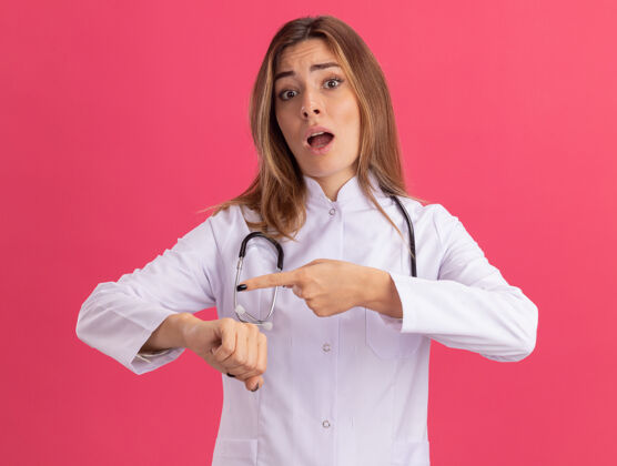 手腕年轻的女医生穿着医用长袍 听诊器显示腕表的手势 隔离在粉红色的墙上长袍女性医生