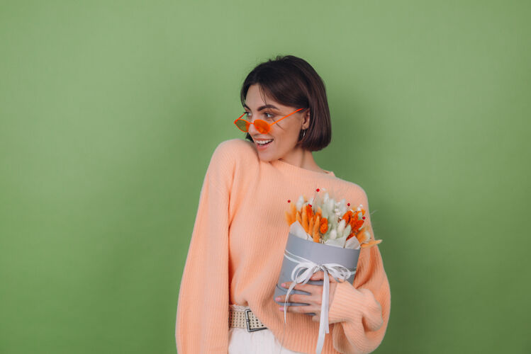 3月8日年轻女子穿着休闲的桃色毛衣隔离在绿橄榄墙上捧着橘白色的花盒组成的棉花 团花 小麦和紫罗兰为礼物开心惊喜黑麦快乐干花