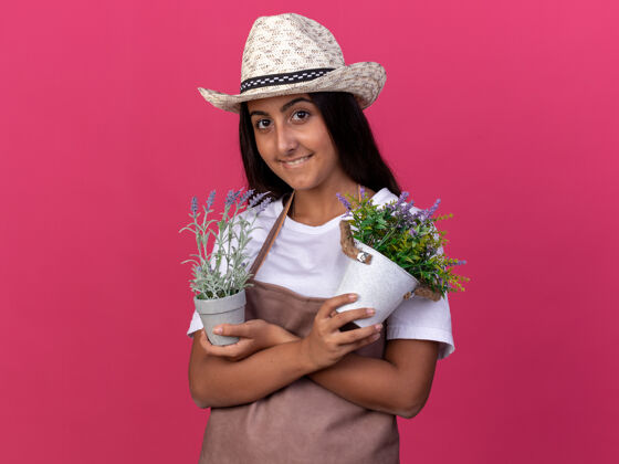 锅穿着围裙 戴着夏帽 手里拿着盆栽植物的快乐的年轻园丁女孩站在粉红色的墙上欢快地笑着帽子围裙花园