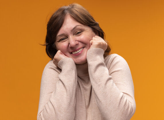 年龄幸福快乐的中年妇女 穿着米色高领毛衣 双手放在脸颊上 站在橙色的墙上中间女人脸颊